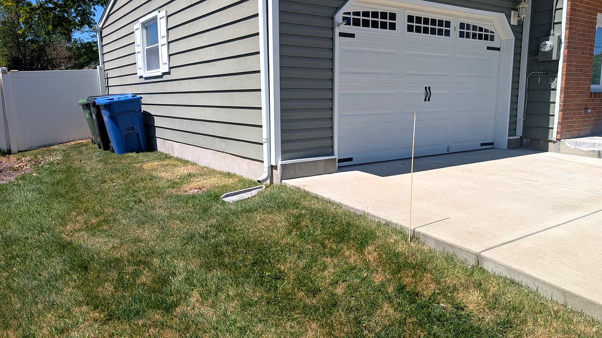Before: trash cans sitting on bare, sloped grass beside the garage with no pad