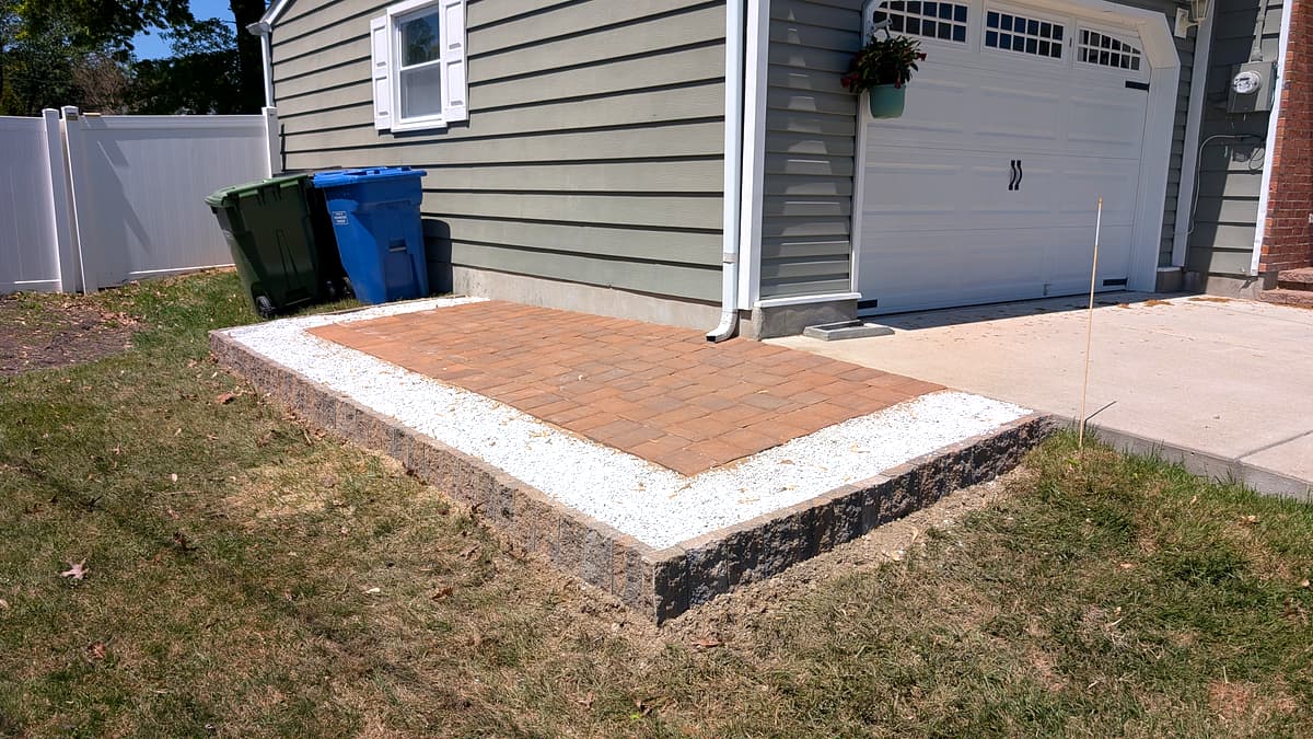 After: elevated paver pad with retaining wall and white stone border for trash cans